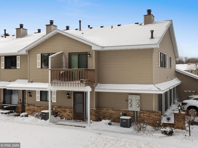 snow covered back of property with central AC, brick siding, a chimney, and a balcony