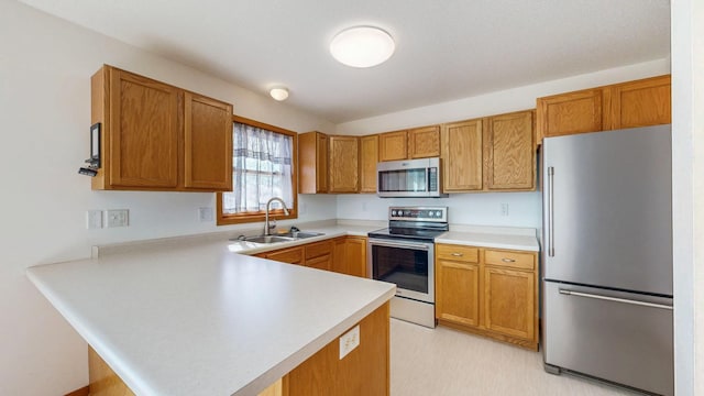 kitchen featuring brown cabinetry, a peninsula, a sink, light countertops, and appliances with stainless steel finishes