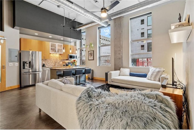 living area with a towering ceiling, a ceiling fan, finished concrete floors, electric panel, and baseboards