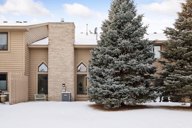 snow covered back of property with central AC, brick siding, and a chimney