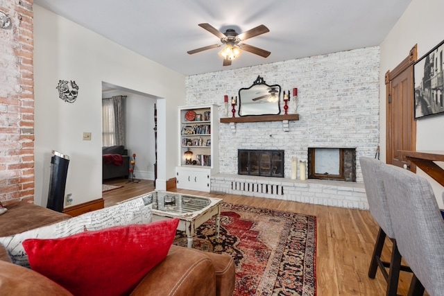 living room featuring wood finished floors, brick wall, a fireplace, baseboards, and ceiling fan