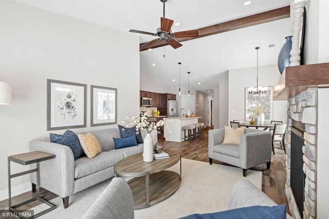 living area featuring high vaulted ceiling, a stone fireplace, ceiling fan with notable chandelier, wood finished floors, and beamed ceiling