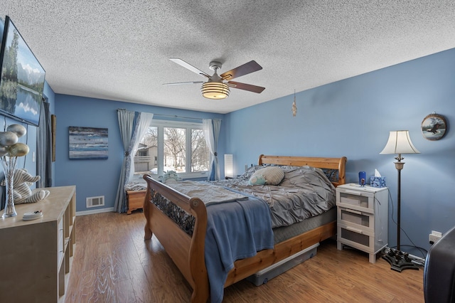 bedroom with visible vents, a textured ceiling, wood finished floors, baseboards, and ceiling fan