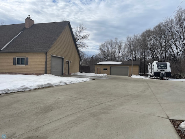 snow covered property featuring a detached garage, an outdoor structure, a chimney, and a shingled roof