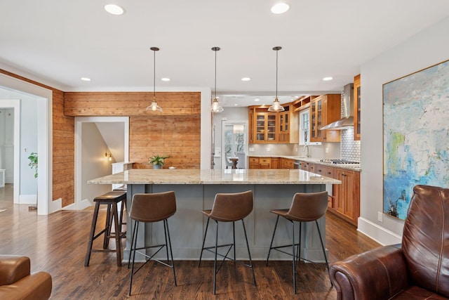 kitchen with dark wood-type flooring, wall chimney range hood, brown cabinets, and stainless steel gas cooktop