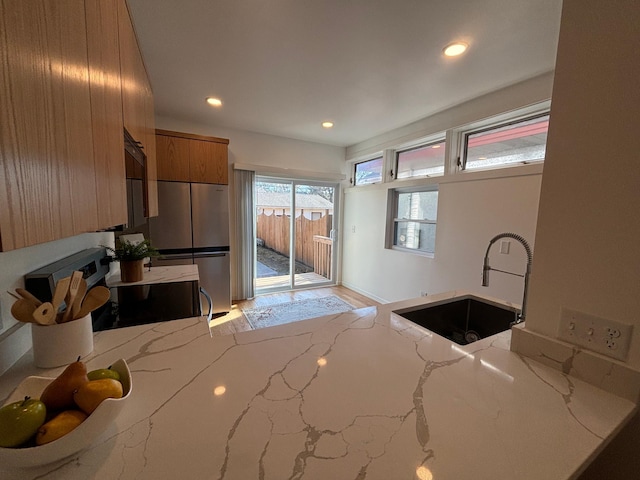 kitchen with electric range, brown cabinetry, freestanding refrigerator, light stone countertops, and a sink