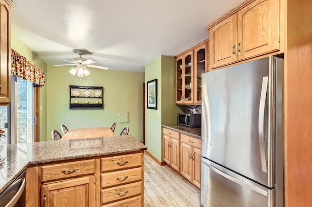 kitchen with ceiling fan, glass insert cabinets, appliances with stainless steel finishes, light stone countertops, and wood finish floors