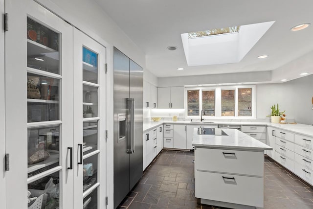 kitchen with a skylight, stone tile floors, built in refrigerator, light stone countertops, and a sink