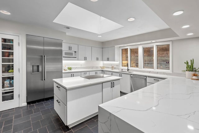 kitchen featuring a skylight, appliances with stainless steel finishes, a kitchen island, a sink, and light stone countertops