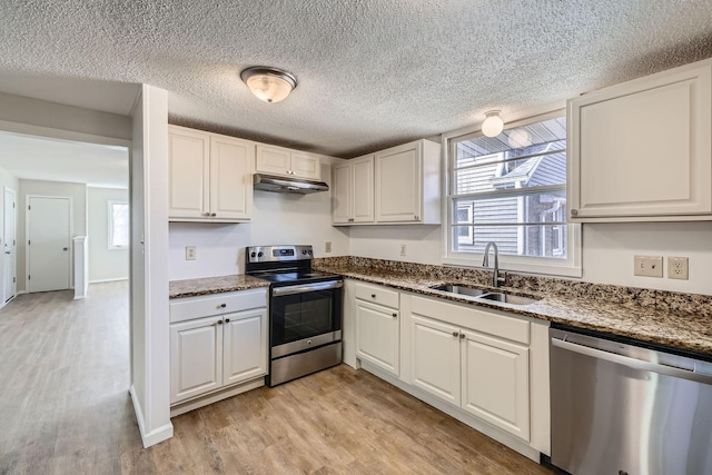 kitchen with dark stone counters, light wood-style floors, appliances with stainless steel finishes, under cabinet range hood, and a sink