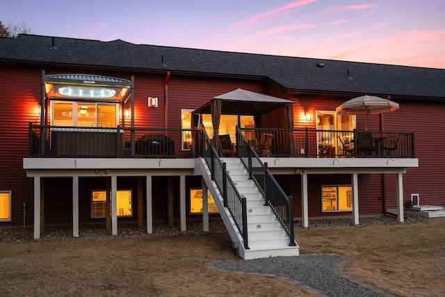 back of house at dusk featuring stairway, a lawn, a wooden deck, and roof with shingles