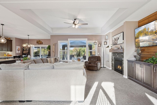 living area with baseboards, light carpet, a glass covered fireplace, a raised ceiling, and a ceiling fan