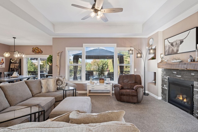 living area featuring carpet, baseboards, a glass covered fireplace, a raised ceiling, and ceiling fan with notable chandelier