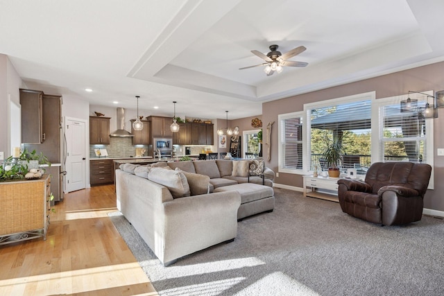 living room with a raised ceiling, ceiling fan with notable chandelier, light wood-type flooring, and baseboards