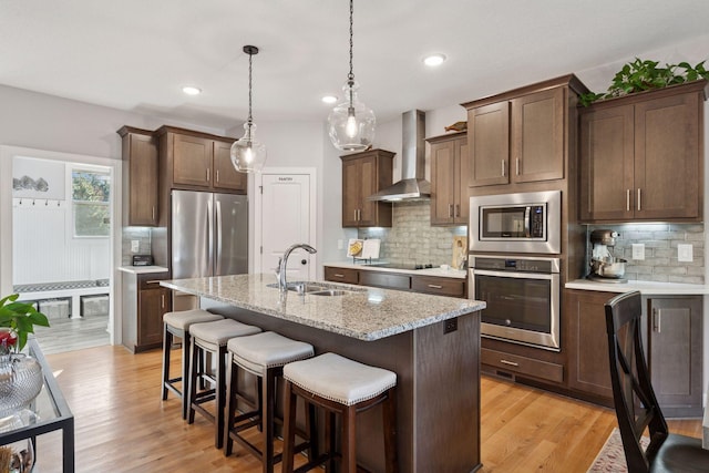 kitchen featuring light wood finished floors, appliances with stainless steel finishes, a kitchen breakfast bar, wall chimney exhaust hood, and a sink