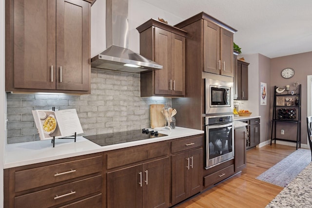 kitchen with light wood-type flooring, light countertops, appliances with stainless steel finishes, wall chimney range hood, and tasteful backsplash