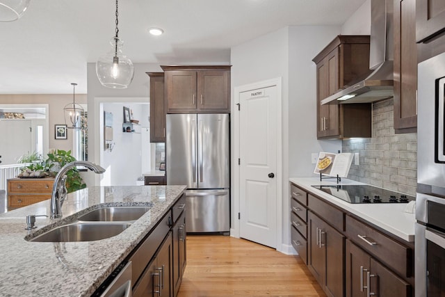 kitchen with light wood finished floors, a sink, appliances with stainless steel finishes, wall chimney range hood, and decorative light fixtures