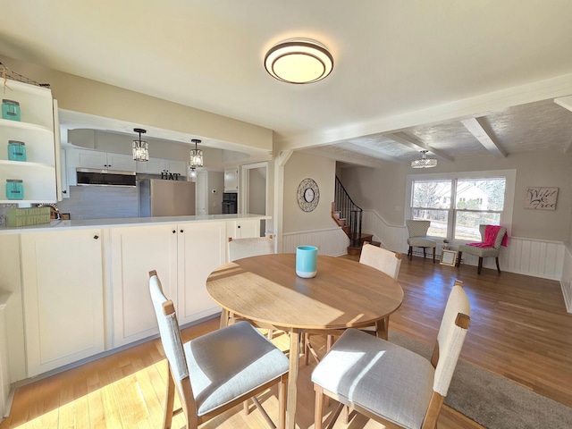 dining area featuring light wood-type flooring, a wainscoted wall, stairway, and beamed ceiling