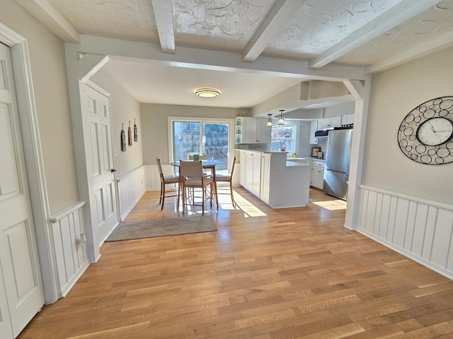 unfurnished dining area featuring light wood-type flooring, a textured ceiling, beamed ceiling, and wainscoting