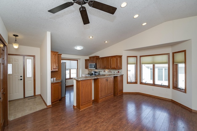 kitchen featuring brown cabinets, dark wood-style floors, stainless steel appliances, a peninsula, and lofted ceiling