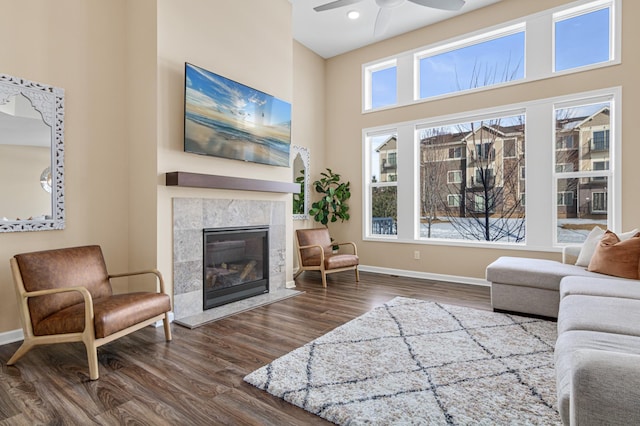 living area featuring a high ceiling, dark wood-style flooring, a ceiling fan, baseboards, and a tiled fireplace