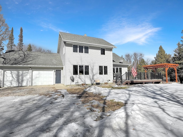 snow covered property with a shingled roof, an attached garage, a wooden deck, and a pergola