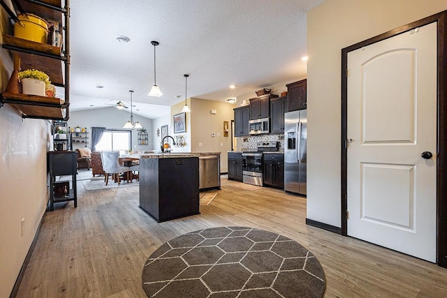 kitchen with light wood finished floors, lofted ceiling, appliances with stainless steel finishes, a kitchen island with sink, and light countertops