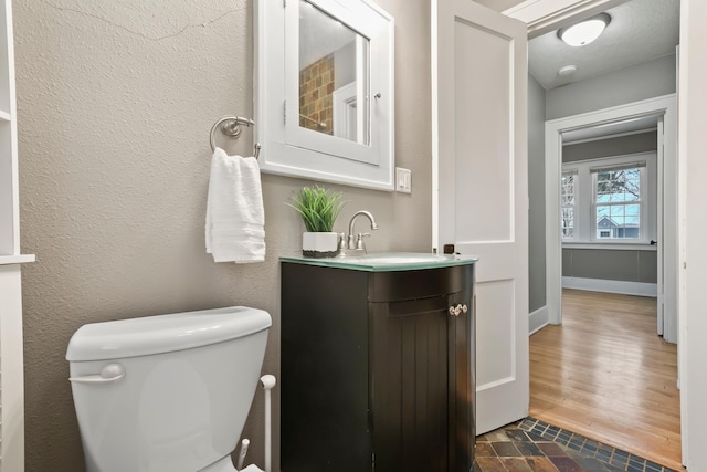 half bathroom with toilet, wood finished floors, baseboards, vanity, and a textured wall