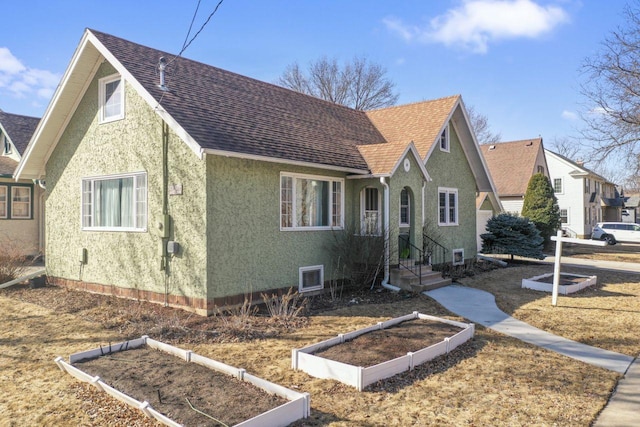 view of front of house with stucco siding, a shingled roof, and a vegetable garden