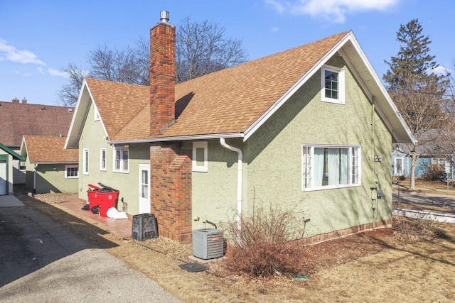 rear view of house featuring stucco siding, cooling unit, a chimney, and a shingled roof