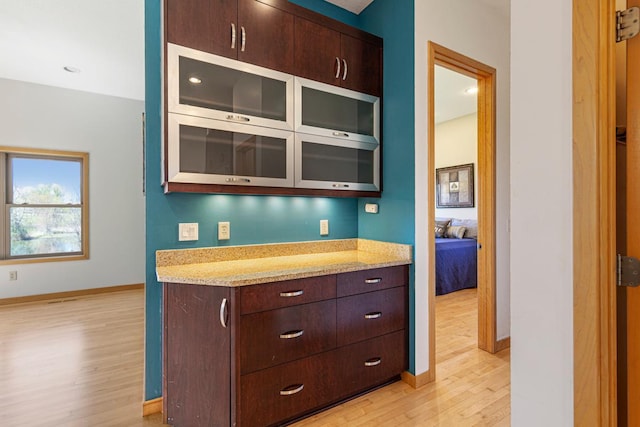 kitchen featuring light wood finished floors, baseboards, dark brown cabinets, glass insert cabinets, and light stone countertops