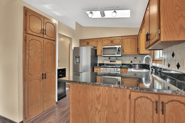 kitchen with a sink, backsplash, stainless steel appliances, brown cabinetry, and vaulted ceiling