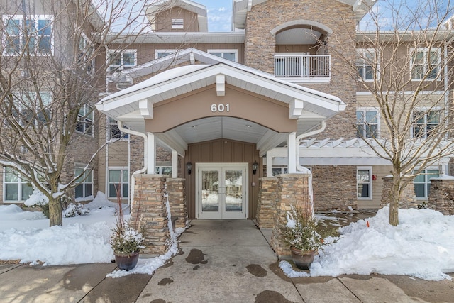 snow covered property entrance featuring board and batten siding, french doors, and stone siding