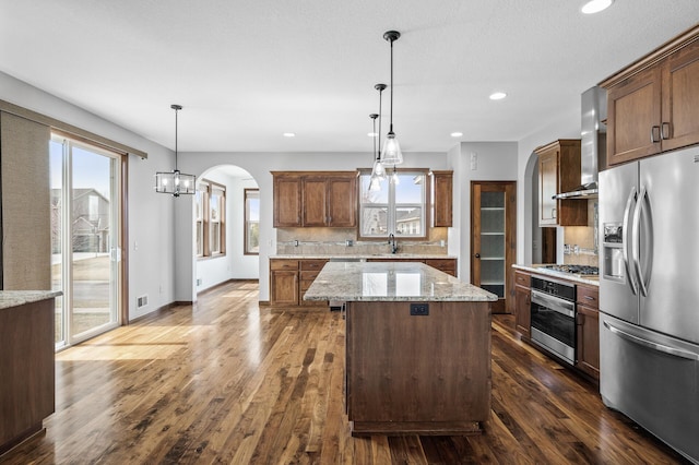kitchen featuring a center island, arched walkways, wall chimney exhaust hood, stainless steel appliances, and decorative backsplash