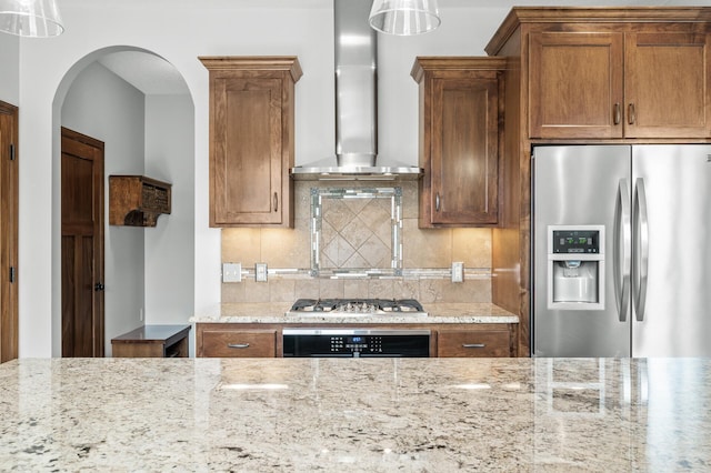 kitchen with tasteful backsplash, wall chimney exhaust hood, brown cabinets, stainless steel appliances, and light stone counters