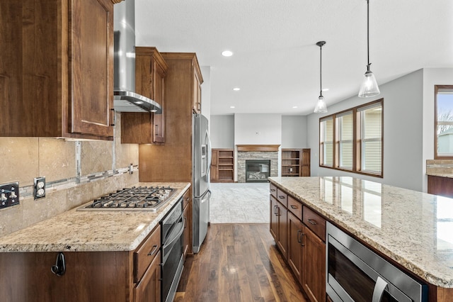 kitchen with a fireplace, wall chimney range hood, appliances with stainless steel finishes, dark wood-style flooring, and open floor plan