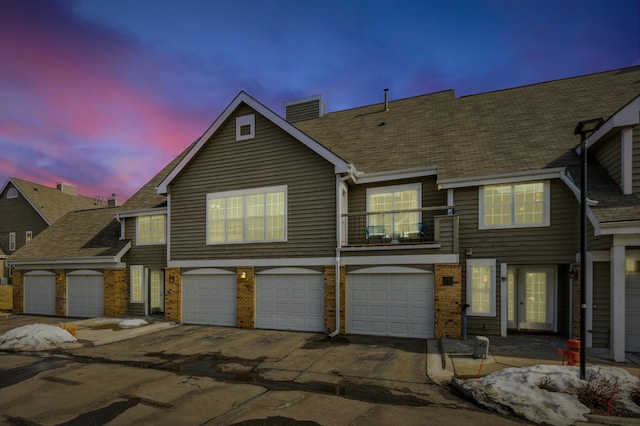 view of property with driveway, a balcony, an attached garage, brick siding, and a chimney