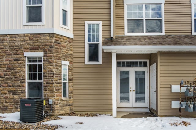 snow covered property entrance with central air condition unit, french doors, roof with shingles, and stone siding