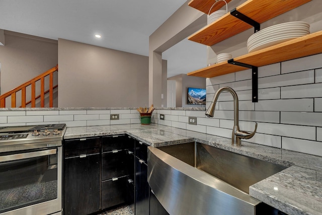 kitchen featuring open shelves, backsplash, stainless steel gas stove, a sink, and dark cabinetry