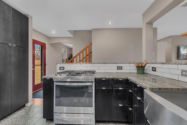 kitchen featuring light stone counters, tasteful backsplash, stainless steel gas stove, dark cabinets, and a peninsula