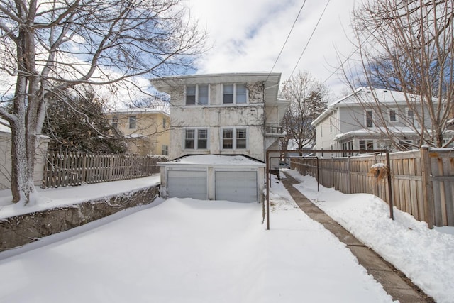 snow covered back of property with a garage, a fenced backyard, an outdoor structure, and stucco siding