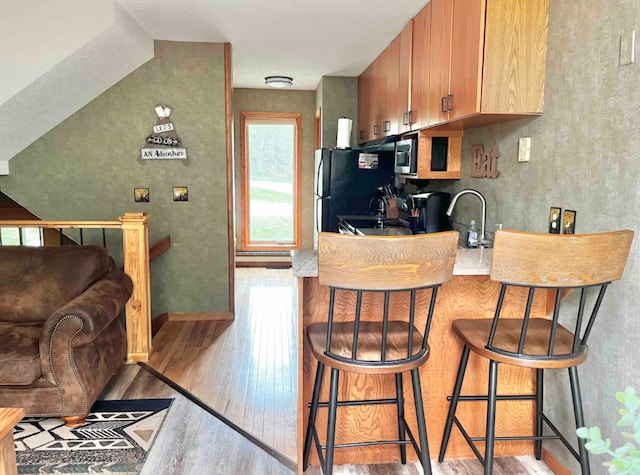 kitchen featuring a breakfast bar, stainless steel microwave, freestanding refrigerator, a sink, and wood finished floors