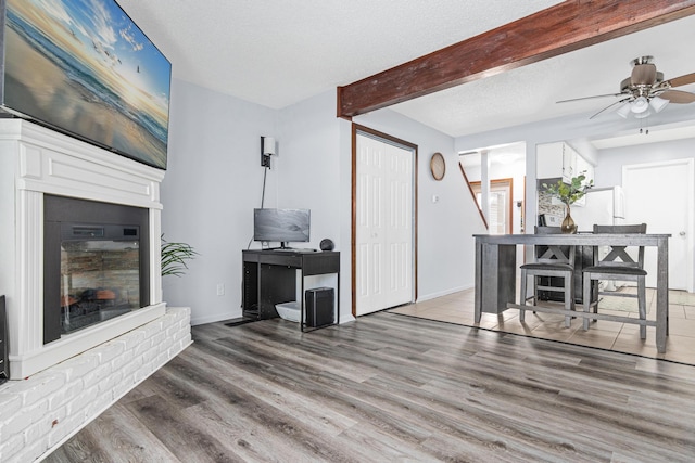 living room with a textured ceiling, wood finished floors, baseboards, a brick fireplace, and beam ceiling