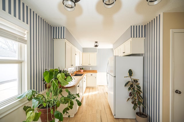 kitchen with light wood finished floors, wallpapered walls, freestanding refrigerator, white cabinets, and a sink