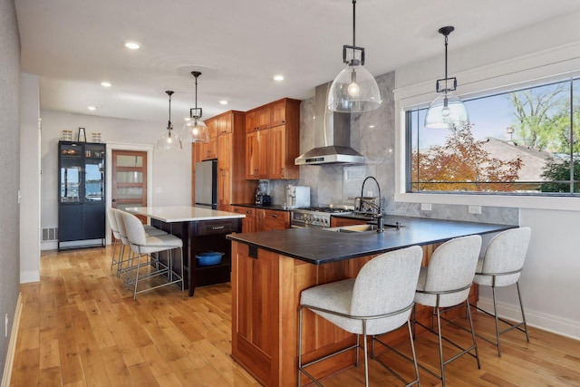 kitchen featuring brown cabinetry, light wood-style flooring, a kitchen breakfast bar, wall chimney range hood, and backsplash