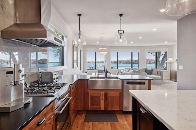 kitchen featuring light wood-style flooring, light stone countertops, stainless steel appliances, wall chimney range hood, and a sink