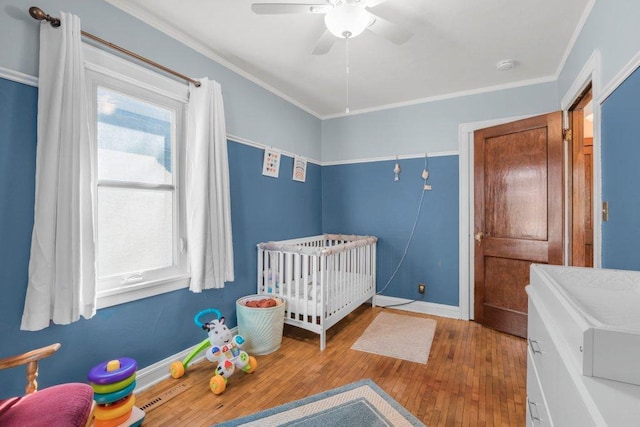 bedroom featuring baseboards, ceiling fan, a crib, wood-type flooring, and crown molding