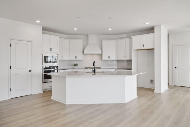 kitchen with decorative backsplash, light wood-style flooring, appliances with stainless steel finishes, a kitchen island with sink, and premium range hood