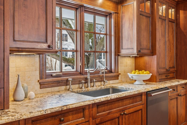 kitchen with backsplash, glass insert cabinets, light stone counters, brown cabinetry, and a sink