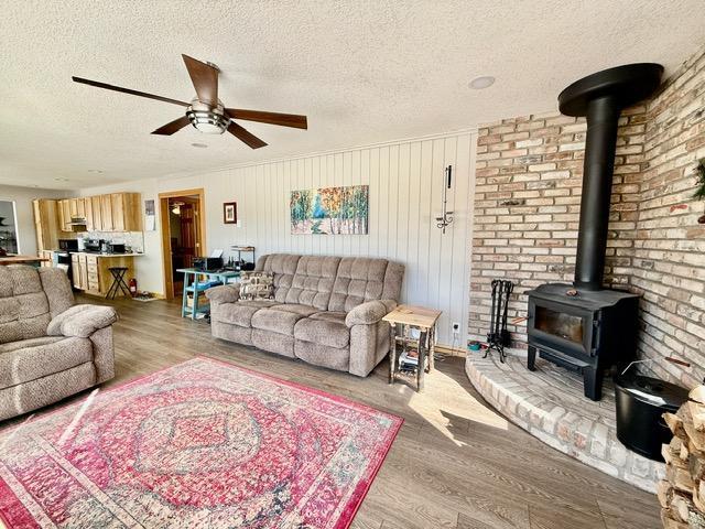 living area with ceiling fan, a textured ceiling, wood finished floors, and a wood stove